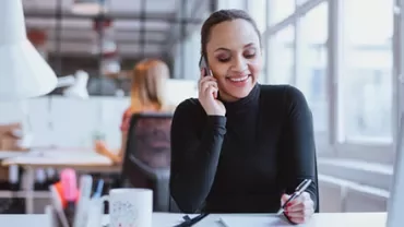 Woman smiling while talking on the phone and taking notes at a desk.