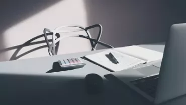A laptop, notebook, pen, and calculator on a desk in sunlight.