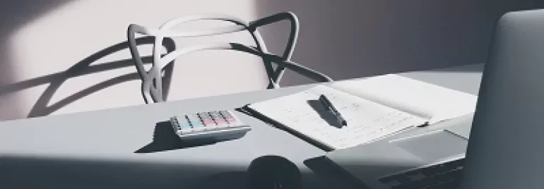Un ordinateur portable, carnet, stylo et calculatrice sur un bureau ensoleillé.